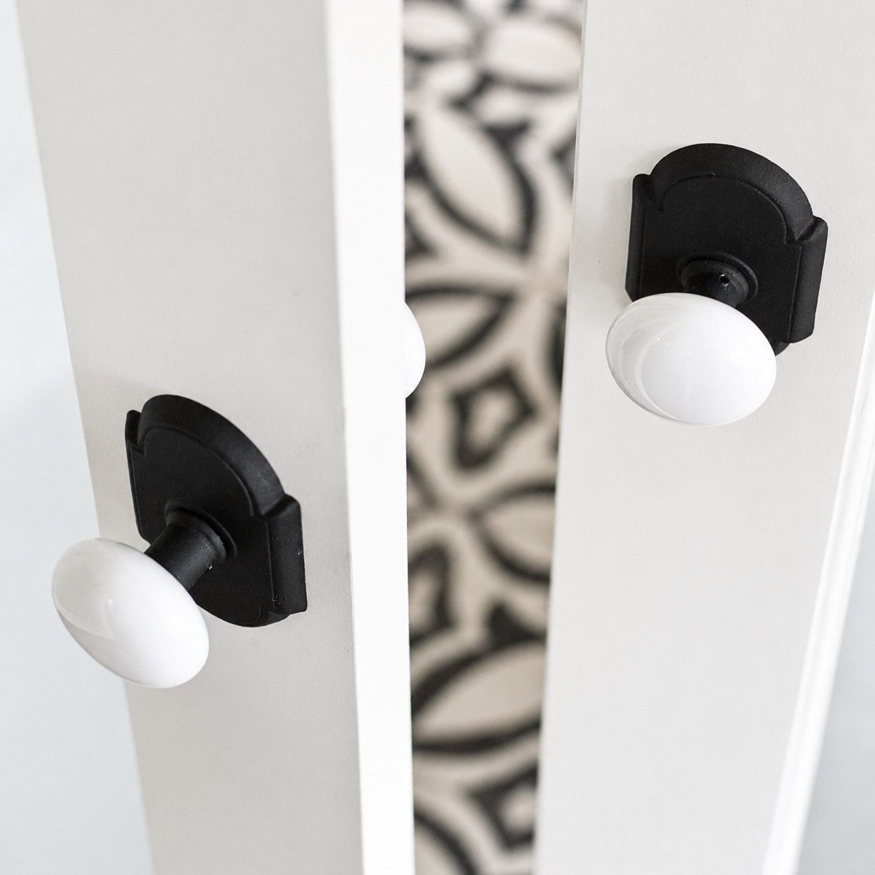 A pair of white oval dummy knobs on matte black arched rosettes, mounted on two white double doors standing slightly open above a patterned black and white tile floor.