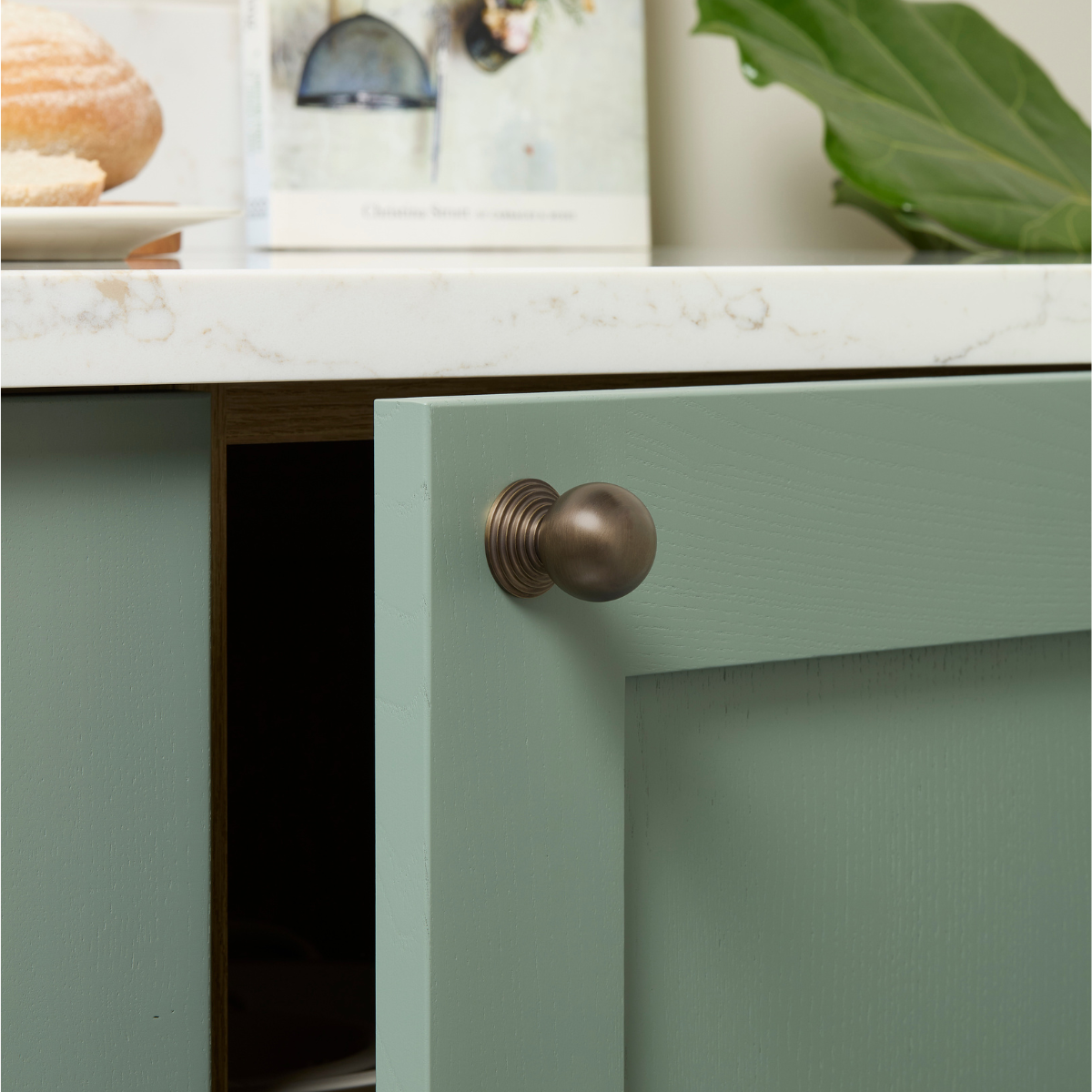 A close-up of a sage green cabinet door with a round, textured bronze knob, set under a white marble countertop with decorative items in the background.