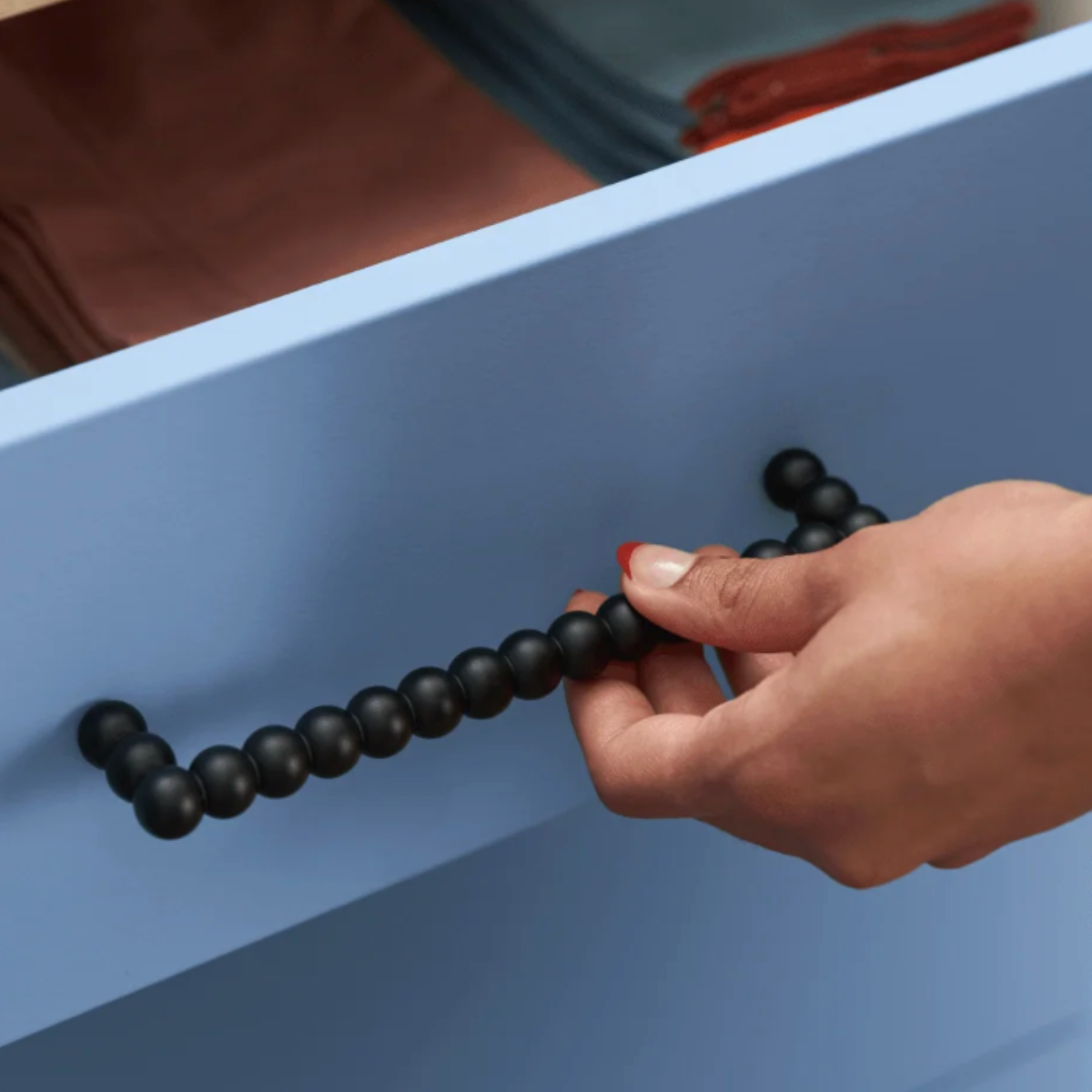 A close-up of a hand with red nail polish pulling a drawer handle made of matte black round beads, set against a light blue drawer front.