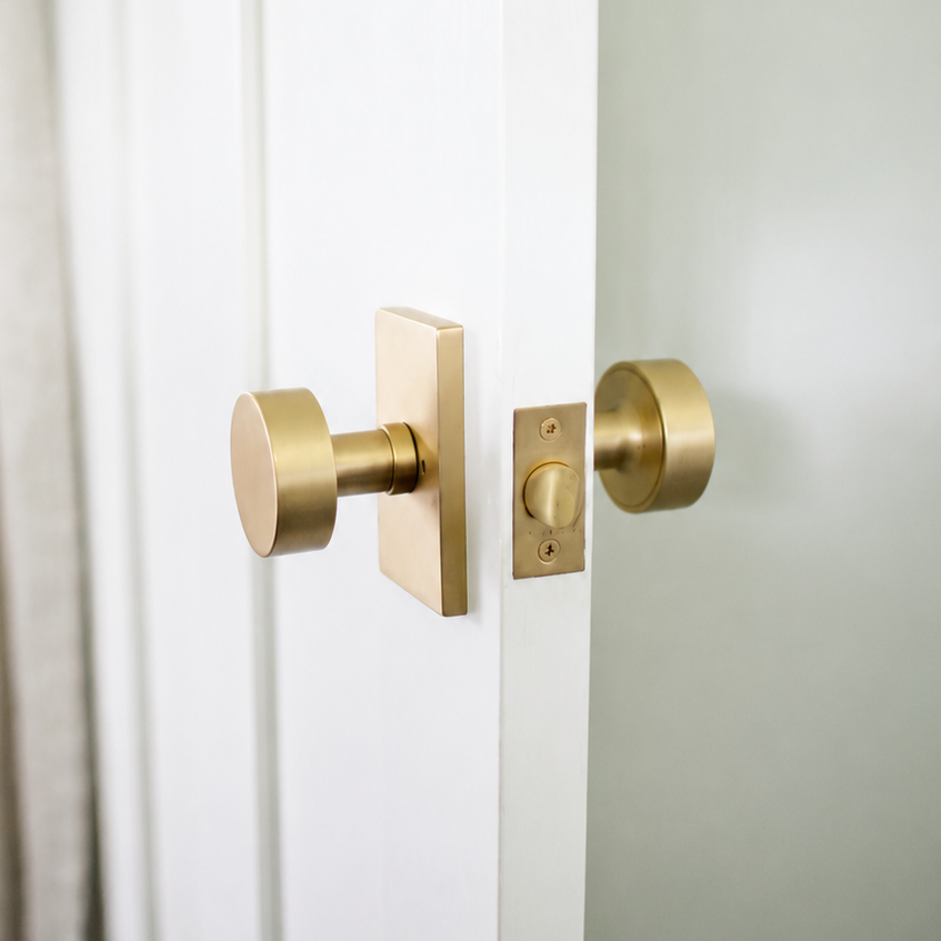 A satin brass privacy knob on a rectangular rosette on a white door, with the brass latch faceplate and turn piece visible along the open edge of the door.