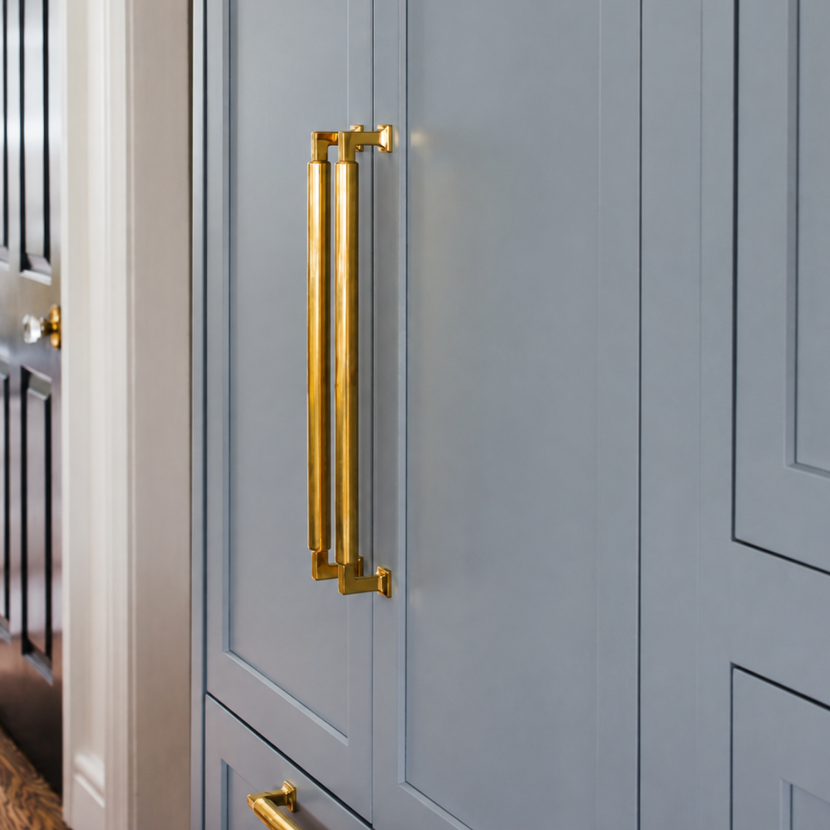 A close-up of a light blue-gray cabinet door featuring a polished gold double-bar pull handle, with part of a matching handle visible on a drawer below.