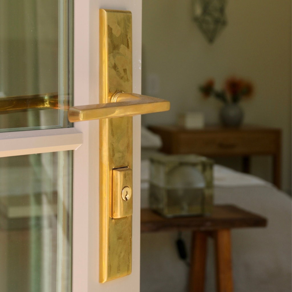 A close-up of a modern brass door handle and lockset mounted on a white door frame, with a softly blurred bedroom background.